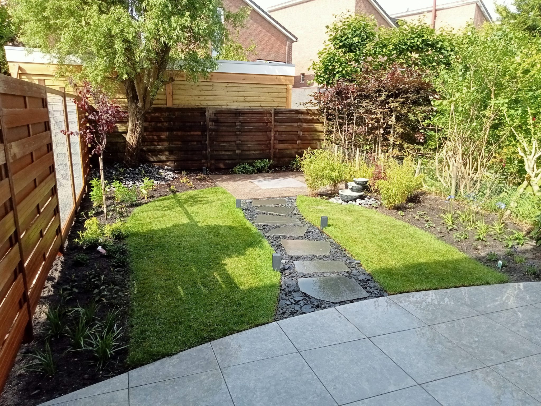 Foto van een organische tuin, genomen vanuit de linkerhoek van het huis op de pagina van tuinonderhoud. Het terras golft door de tuin, gelegd met marmerachtige tegels. Aan weerszijden van de schutting groeien planten, struiken en bomen. In het midden van de tuin ligt een golvend grasveld, omringd door groen. Het einde van de tuin is betegeld, verbonden met natuurlijke stenen, keitjes en tuinlampjes. Achterin staat een grote mediterrane boom. Keko Hoveniers biedt professioneel tuinonderhoud als een Hovenier in voorburg, Nootdorp, Wassenaar, Pijnacker, Zoetermeer, Rijswijk, Berkel en Rodenrijs en omgeving.