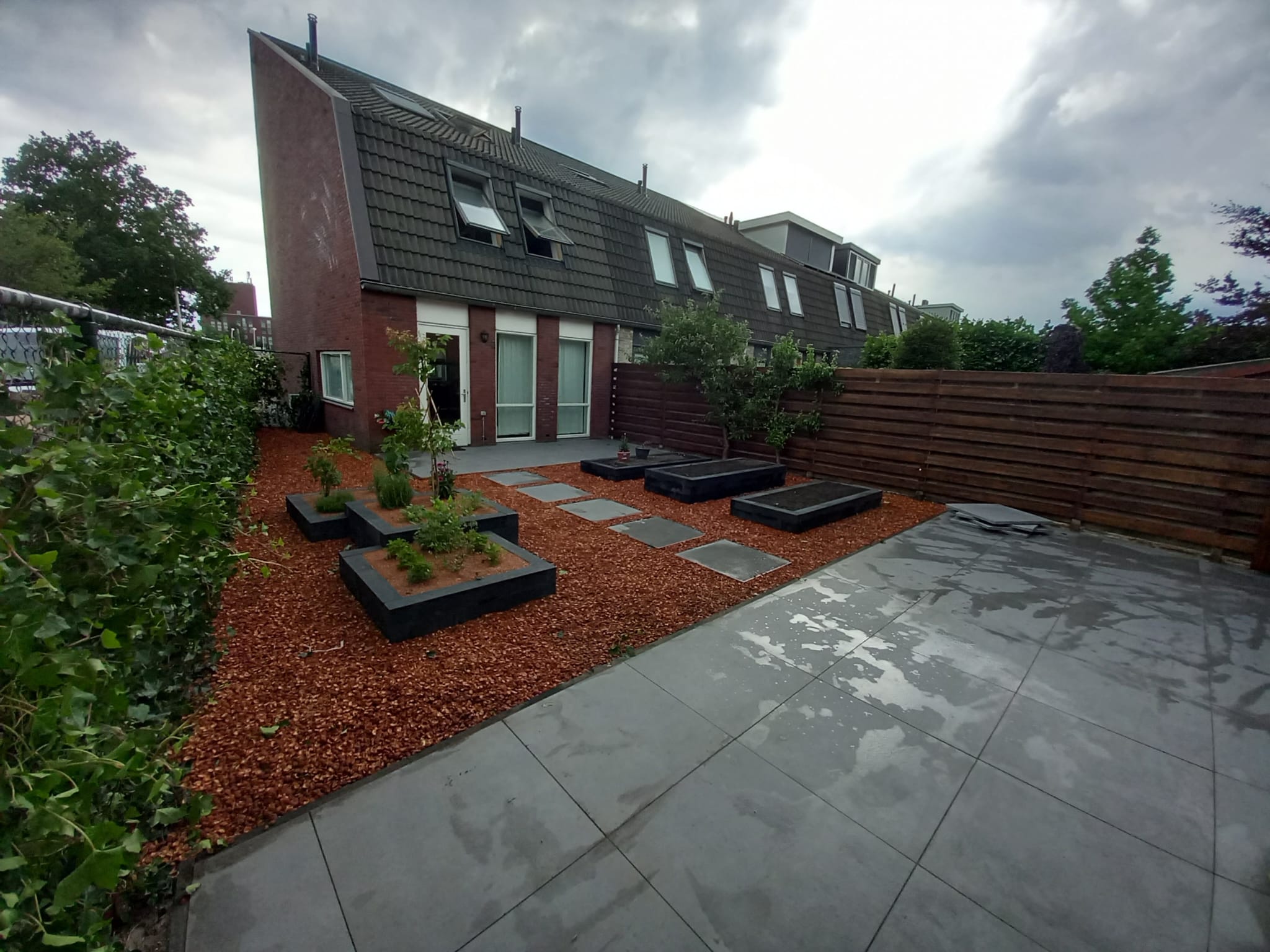 Moderne tuin in Pijnacker, ontworpen en aangelegd door Keko Hoveniers. De tuin begint vanuit de linkerhoek aan het einde van het huis, waarbij grote strakke grijze tegels zijn gebruikt bij de achterdeur en aan het einde van de tuin. De rest van de tuin is bezaaid met boomstronken, terwijl er stenen zijn geplaatst vanaf het huis naar de achterkant van de tuin, dezelfde tegels als elders. In het midden van de tuin zijn meerdere plantenbakken asymmetrisch geplaatst, en langs de tuin is klimop geplant voor privacy langs het ijzeren hek.