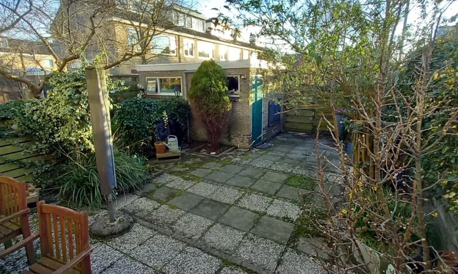 Alt-tekst: "Vóór-foto van een moderne tuin in Nootdorp, voordat Keko Hoveniers aan de slag ging. Deze tuin is ontworpen met oog voor detail, vanuit de rechterhoek van het huis. Het betegelde terras vormt een halfronde oase, omringd door een weelderig perkje met afwisselende struiken en planten. In het hart van de tuin prijken keitjes, terwijl de schutting, schuur en overkapping zijn opgetrokken uit warm oranje hout, met overlappende planken voor een rustieke uitstraling. De schuur is bekroond met een groen dak, bedekt met keitjes en mos, en heeft een subtiele helling voor waterafvoer. Aan het einde van de tuin nodigt een sfeervolle overkapping uit tot ontspanning, met een hoekige lounge set voorzien van comfortabele grijze kussens. Keko Hoveniers biedt professionele tuinaanleg diensten in Nootdorp, Zoetermeer, Wassenaar, Pijnacker, Rijswijk, Voorburg, Berkel en Rodenrijs.